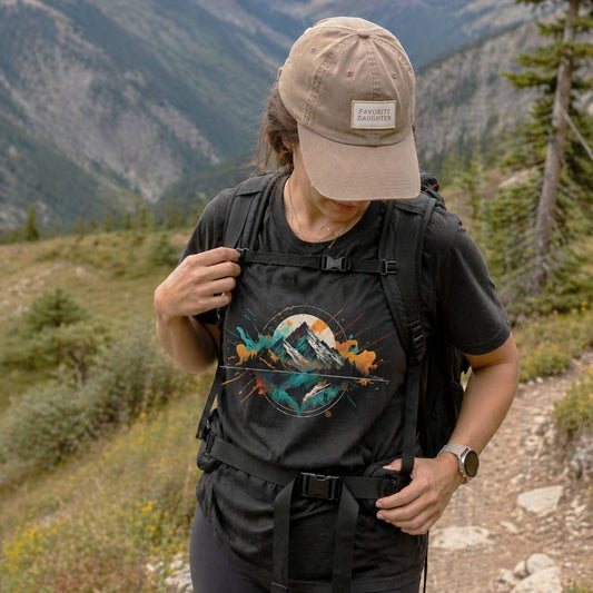 Person hiking with a black backpack featuring a mountain design, wearing a beige cap and black shirt, against a mountainous background.