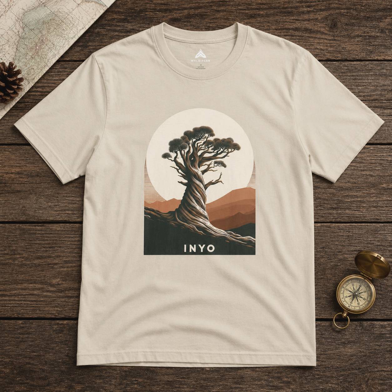 Beige t-shirt with a tree graphic and 'INYO' text on a wooden surface.