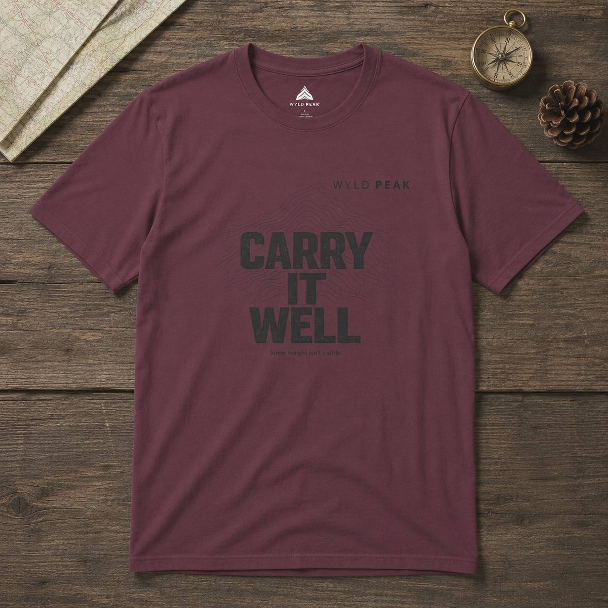 Maroon t-shirt with 'CARRY IT WELL' text on a wooden surface, featuring Wild Peak branding.