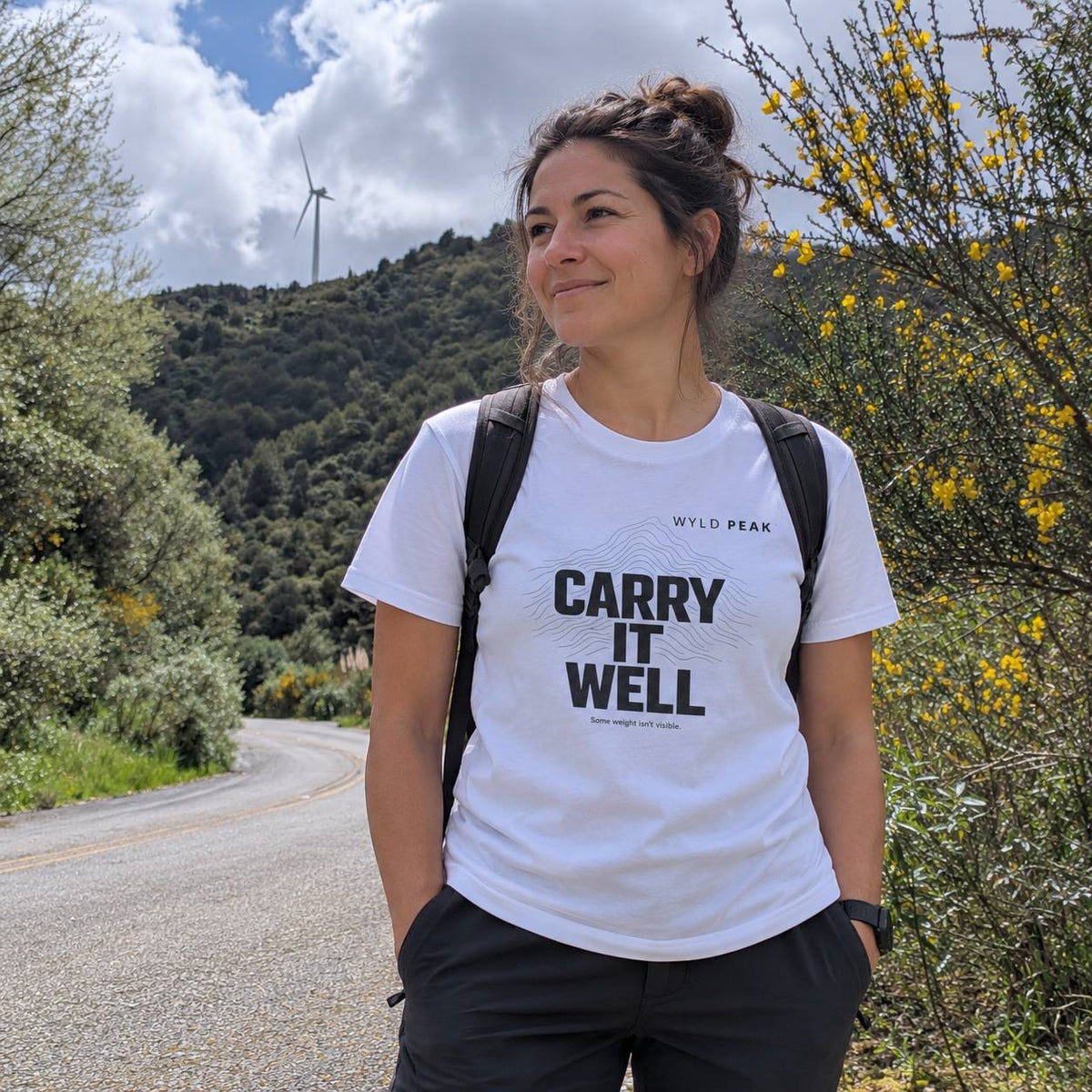 A woman standing outdoors on a road with mountains, trees, and yellow flowers in the background, wearing a white t-shirt with the text 'CARRY IT WELL' and 'WYLO PEAK' printed on the front, along with black pants and a black backpack.
