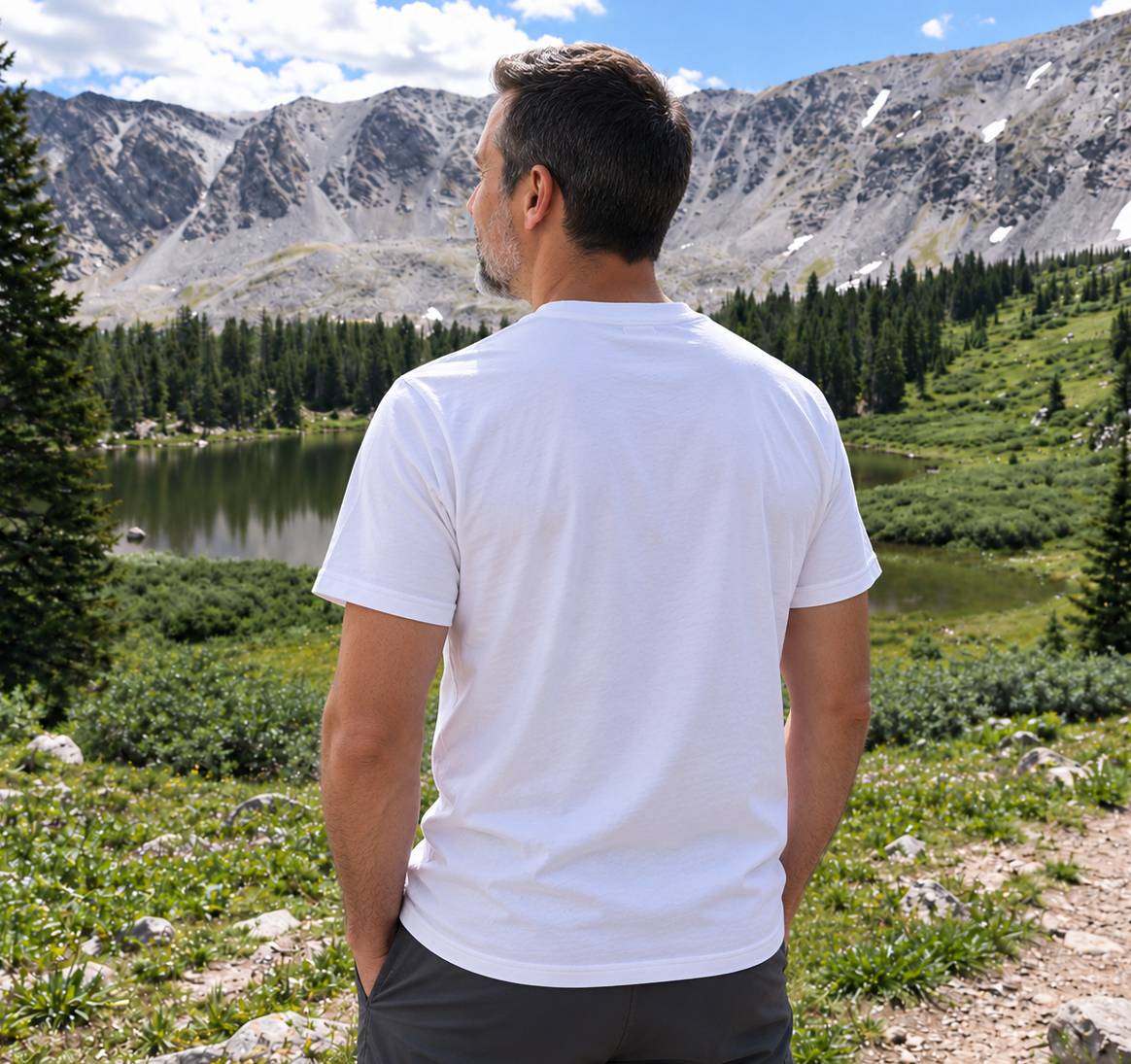 A man viewed from behind wearing a plain white short-sleeve t-shirt and dark gray shorts, standing in a mountain meadow with a lake and pine trees in the background.