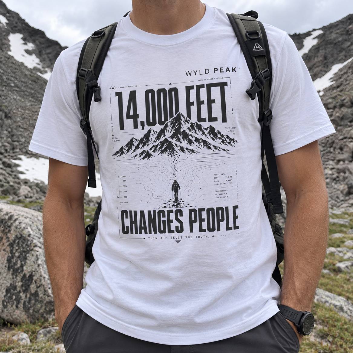 A man wearing a white graphic t-shirt with a black mountain print and the text '14,000 FEET' and 'CHANGES PEOPLE'. The t-shirt features 'WYLD PEAK' branding at the top.