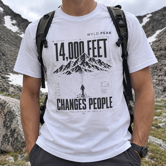 A man wearing a white graphic t-shirt with a black mountain print and the text '14,000 FEET' and 'CHANGES PEOPLE'. The t-shirt features 'WYLD PEAK' branding at the top.