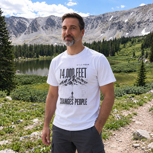 A man wearing a white graphic t-shirt with '14,000 FEET' and 'CHANGES PEOPLE' printed on the front, standing outdoors in a mountain landscape with a lake and pine trees in the background.