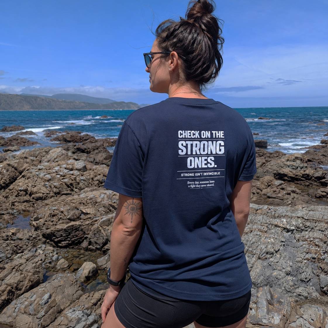 A woman stands on rocky coastal ground facing the ocean, wearing a navy blue T-shirt with bold white text on the back that reads 'CHECK ON THE STRONG ONES.' and smaller supporting text.