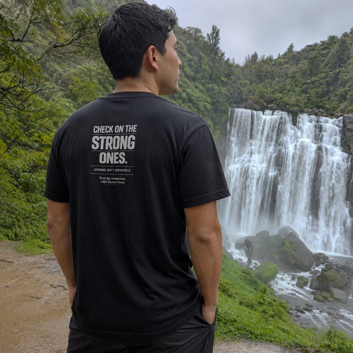 A man stands outdoors in front of a large waterfall, wearing a black T-shirt with white printed text on the back that reads, 'CHECK ON THE STRONG ONES.'.