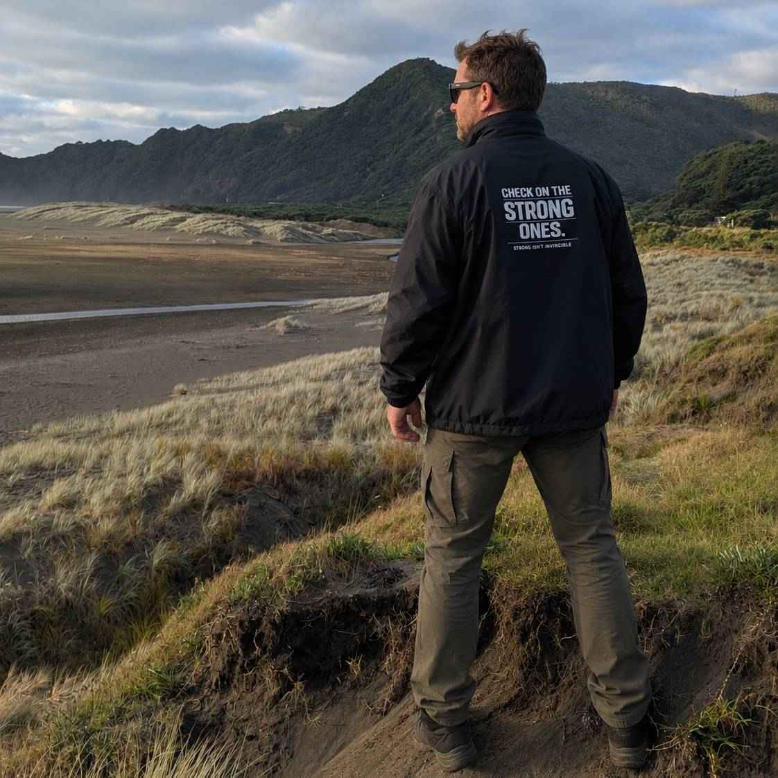 A man wearing a black jacket with the text 'CHECK ON THE STRONG ONES.' on the back stands on a grassy hill overlooking a wide, rugged landscape with mountains and a river.
