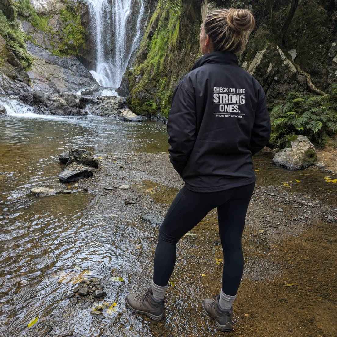 A woman stands in a shallow stream near a waterfall, viewed from behind, wearing a black outdoor jacket with white text that reads 'CHECK ON THE STRONG ONES.' and smaller text below.