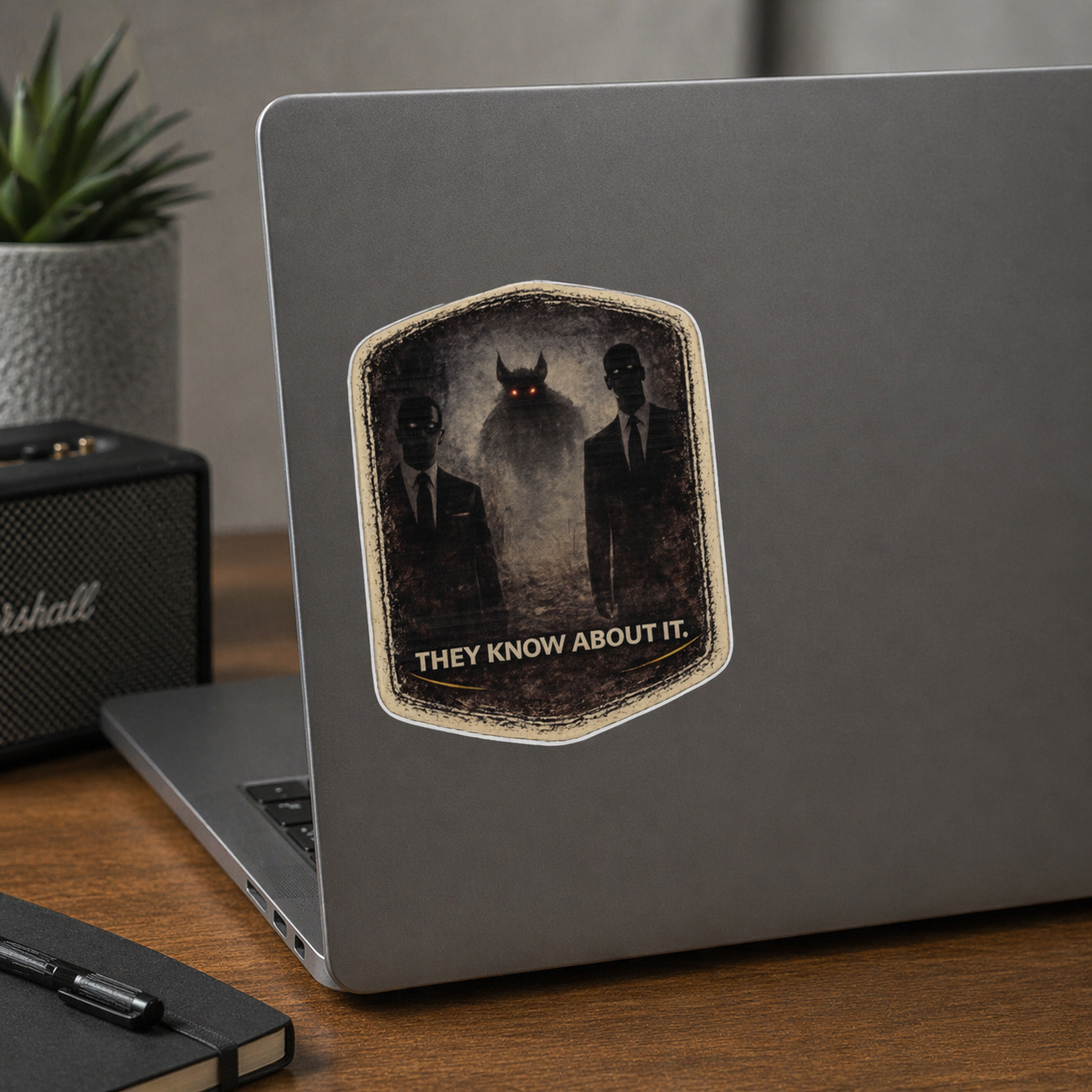 Laptop with a sticker featuring two people and a creature, with text 'THEY KNOW ABOUT IT.' on a wooden desk.