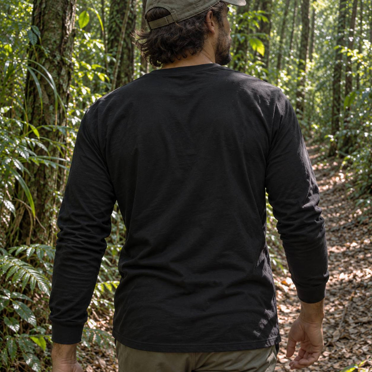 A man wearing a black long-sleeve t-shirt and a khaki cap, viewed from behind as he walks along a forest path.