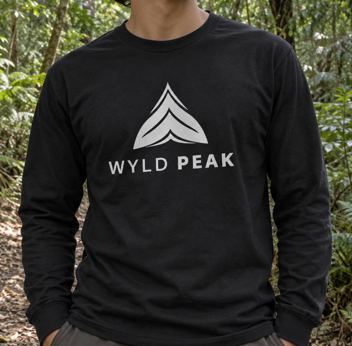 A person wearing a black long-sleeve t-shirt with a white 'WYLD PEAK' logo and chevron graphic on the chest, photographed outdoors in a forest setting.
