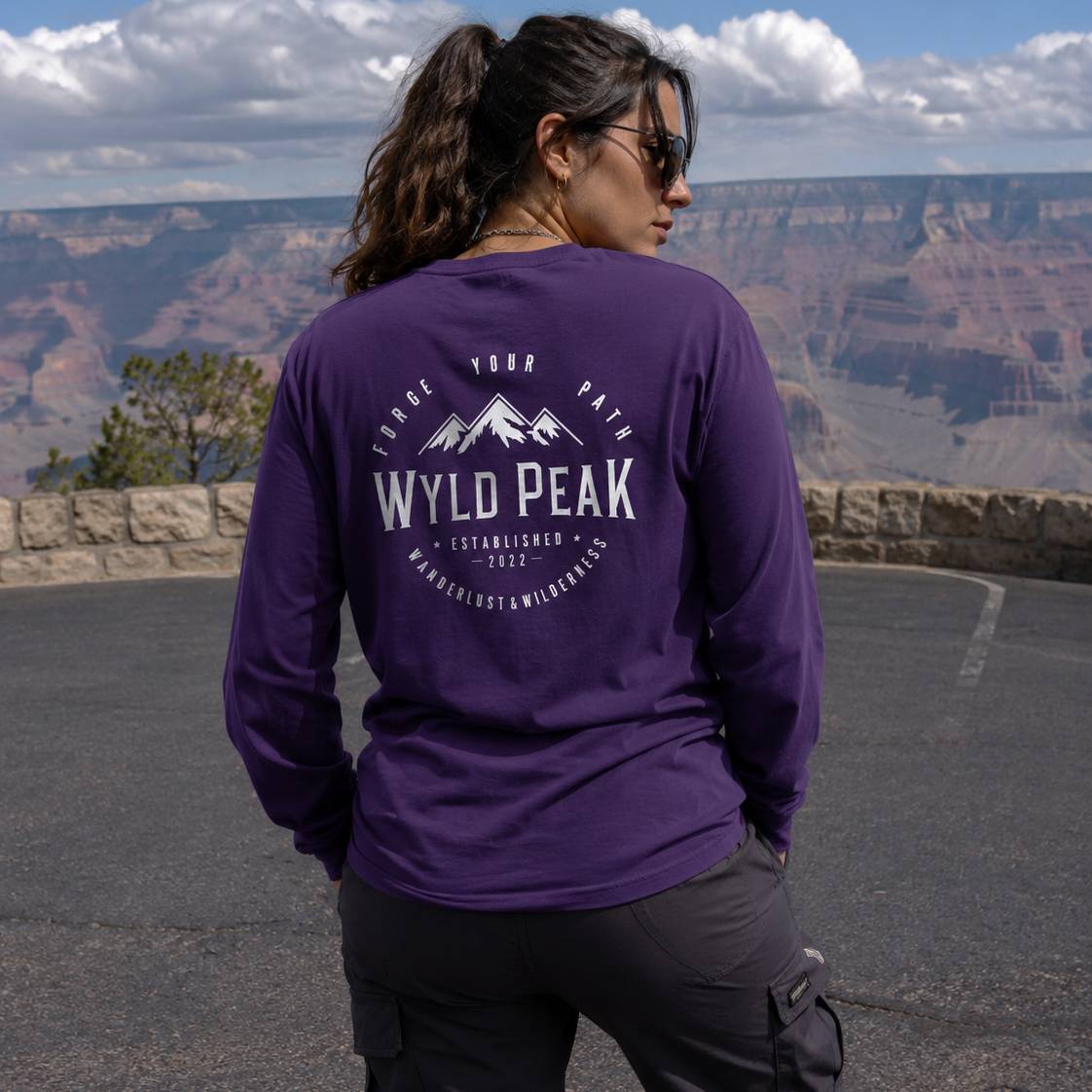 Person wearing a purple 'Wyld Peak' shirt with a scenic view of a canyon.