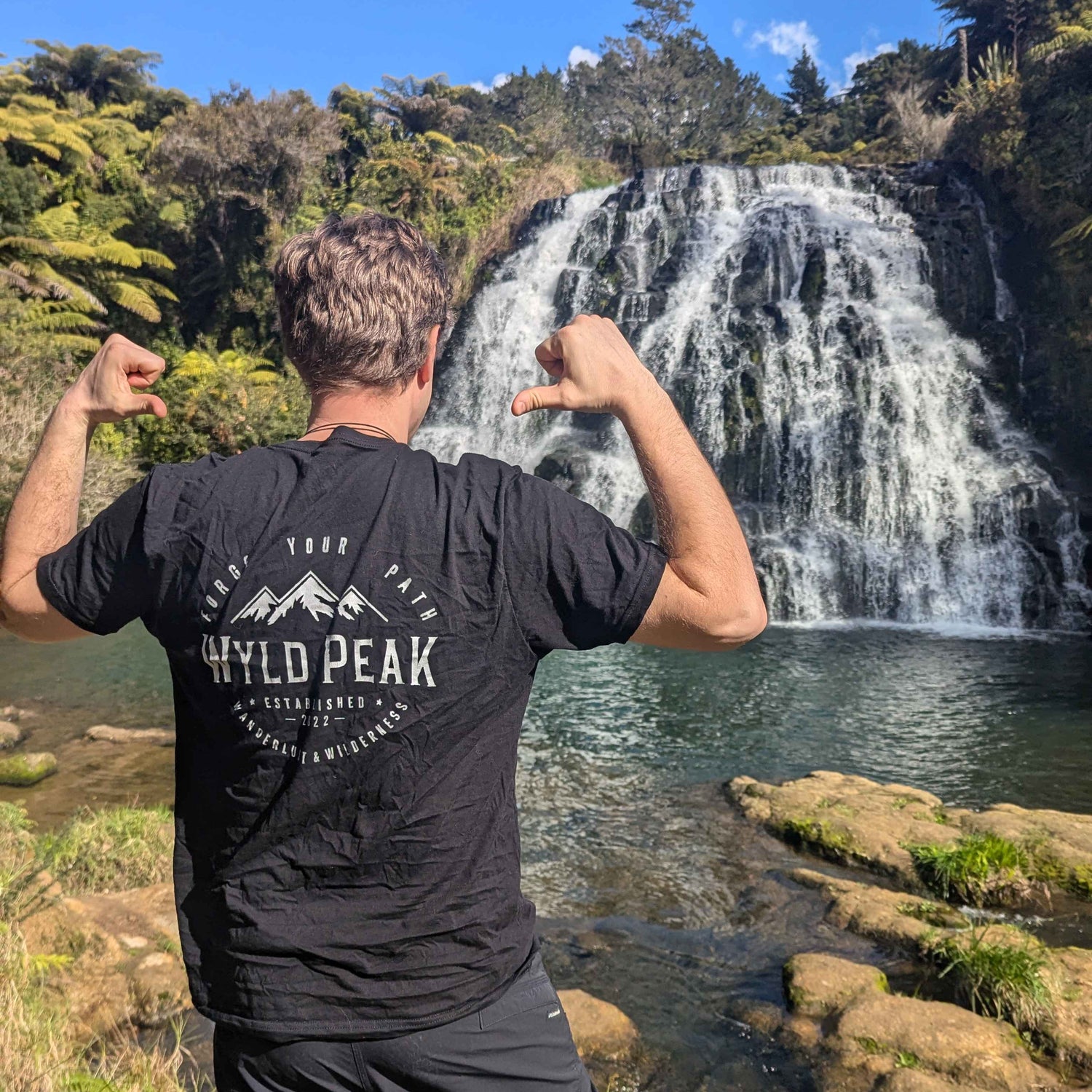 Person flexing muscles in front of a waterfall wearing a black t-shirt with 'Wyld Peak' branding.