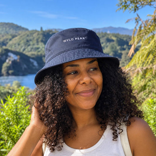A woman outdoors wearing a navy blue bucket hat with 'WYLD PEAK' embroidered in white on the front, with a scenic mountain and lake background.