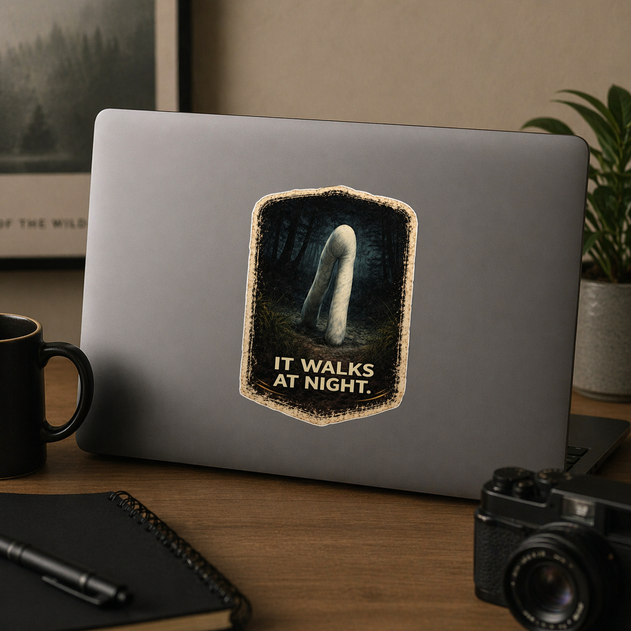 Laptop with a 'It Walks at Night' sticker on a desk with a camera and mug.