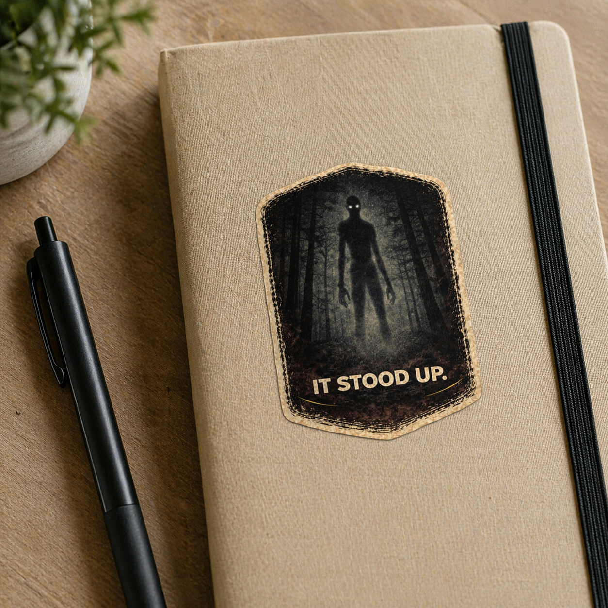 Beige notebook with a black and white illustration of a figure and text 'IT STOOD UP.' on a wooden surface.