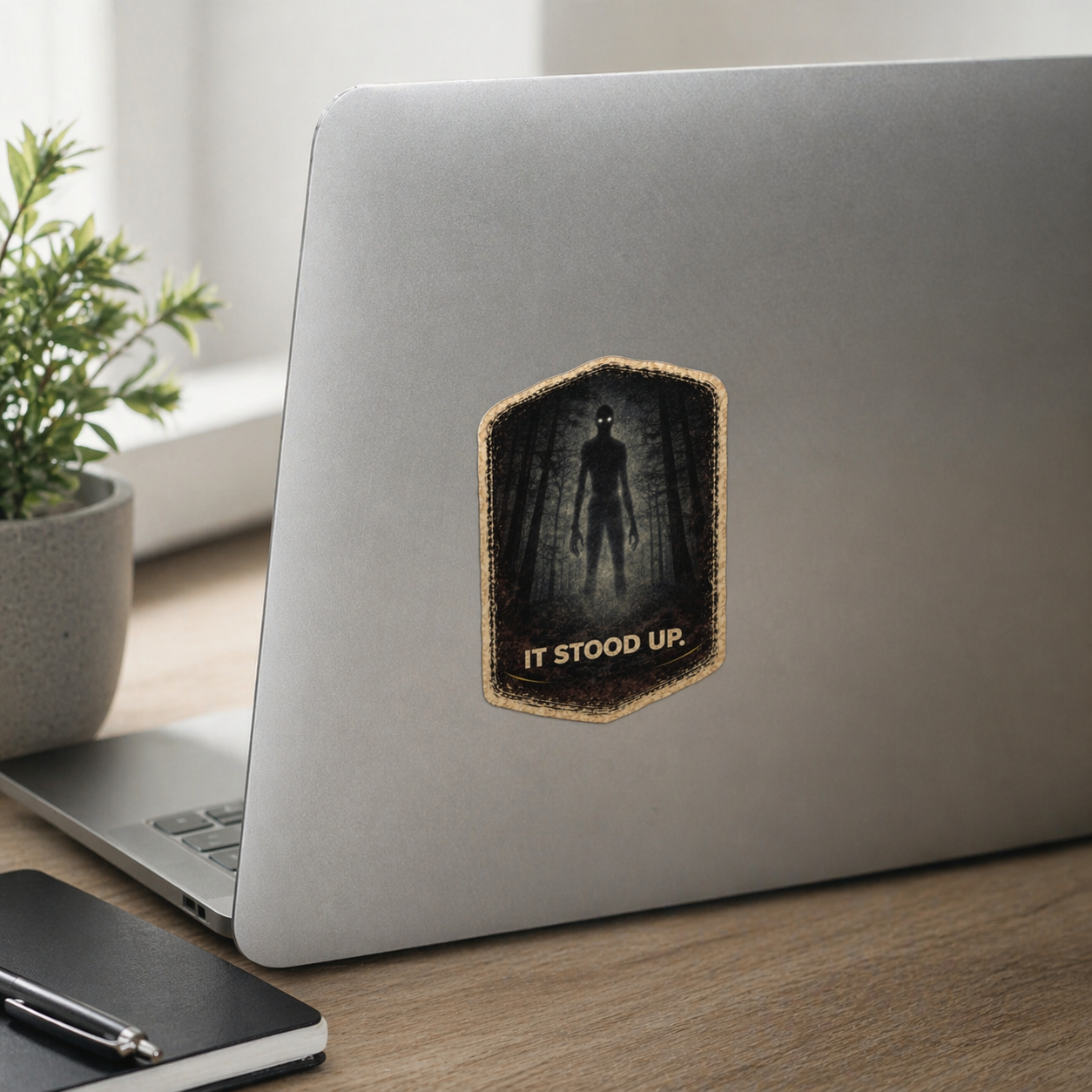 Laptop with a 'It Stood Up' sticker on a wooden desk with a plant in the background