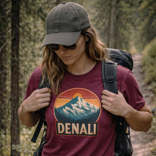 Person hiking in a forest wearing a maroon t-shirt with Denali design and a gray cap.