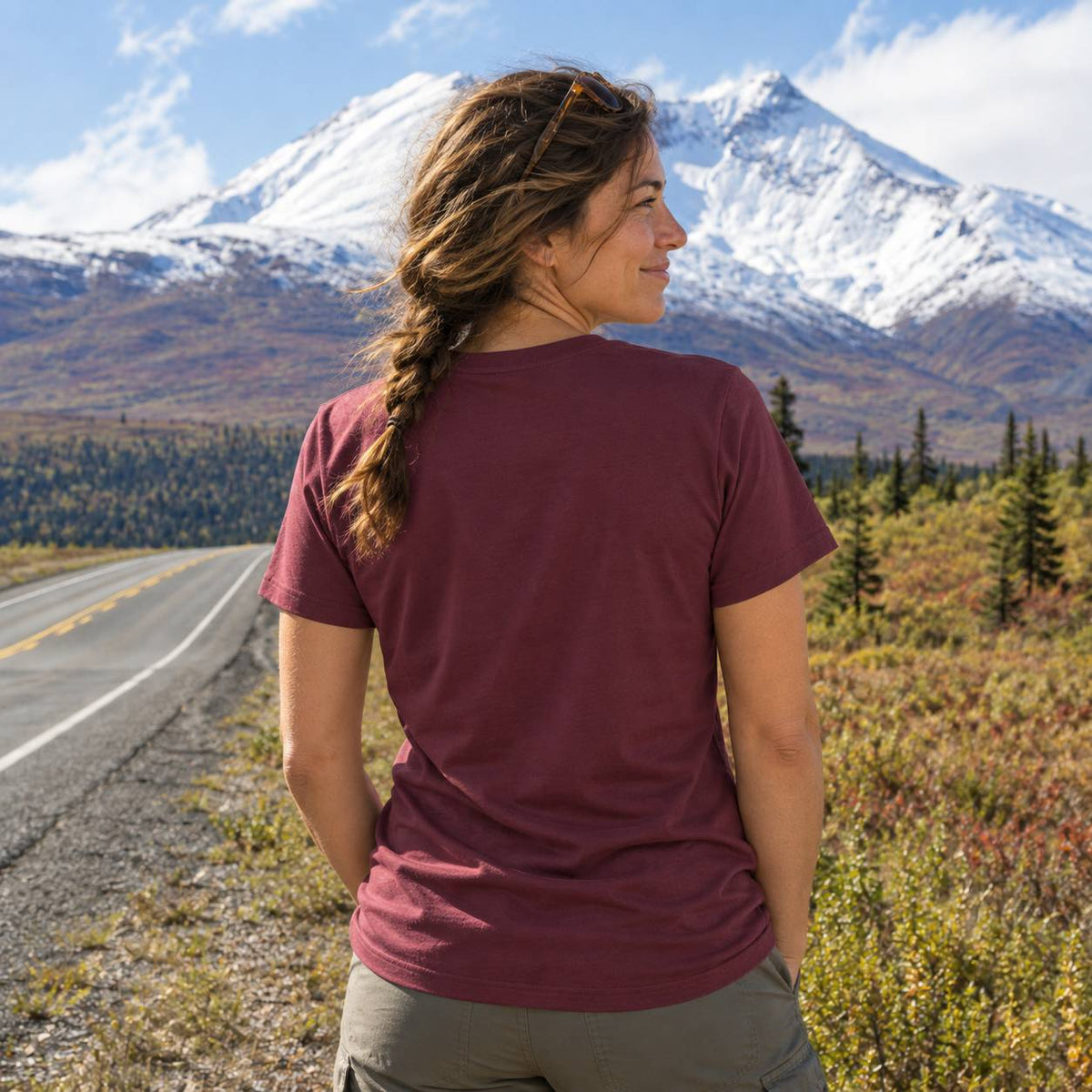 A woman wearing a short-sleeve crewneck t-shirt in burgundy, photographed from behind as she looks toward a snow-capped mountain range on a road trip.
