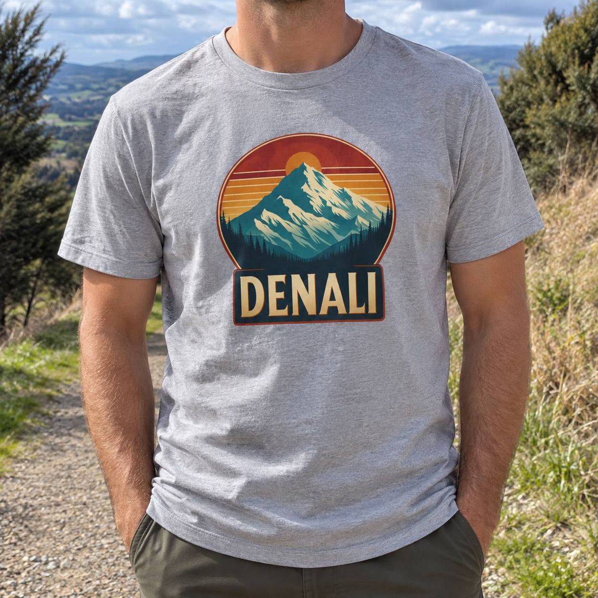 A man wearing a heather gray t-shirt with a retro mountain graphic and the text 'DENALI' on the front, photographed outdoors in a scenic mountain landscape.
