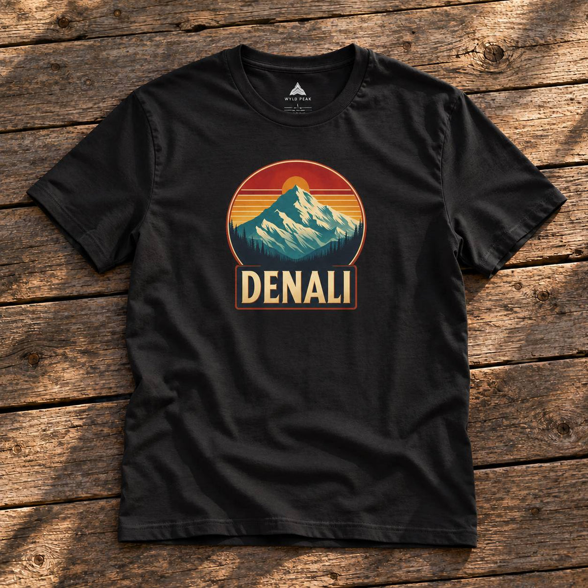 A black short-sleeve t-shirt laid flat on a rustic wooden surface, featuring a retro mountain graphic with the word 'DENALI' below it.