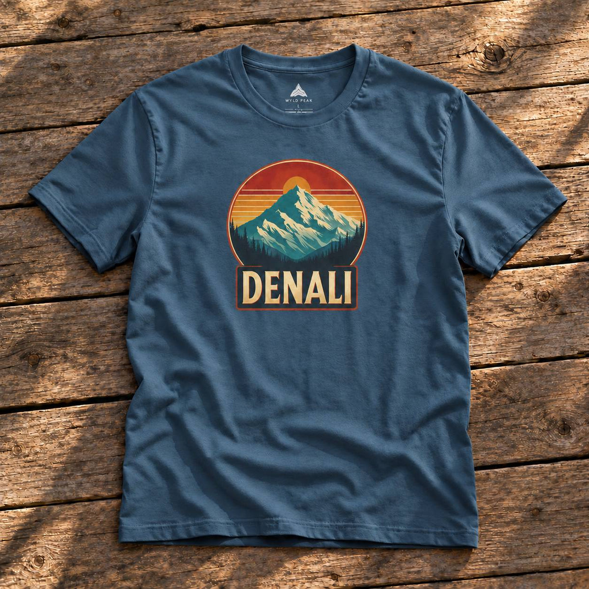 A slate blue crewneck t-shirt with a retro mountain sunset graphic and the word 'DENALI' printed on the chest, laid flat on a rustic wooden plank background.