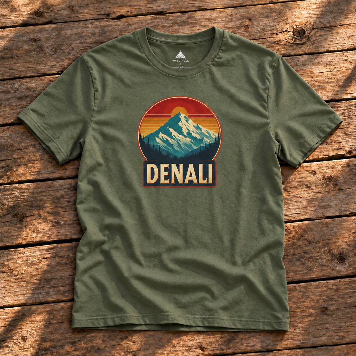 An olive green t-shirt laid flat on a rustic wooden surface, featuring a retro-style mountain graphic with the word 'DENALI' on the chest.