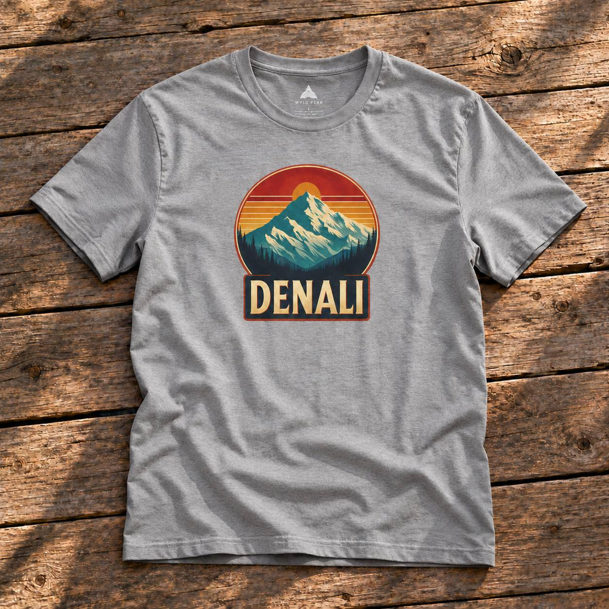 A heather gray short-sleeve t-shirt with a retro mountain sunset graphic and the word 'DENALI' printed on the chest, laid flat on a rustic wooden plank surface.
