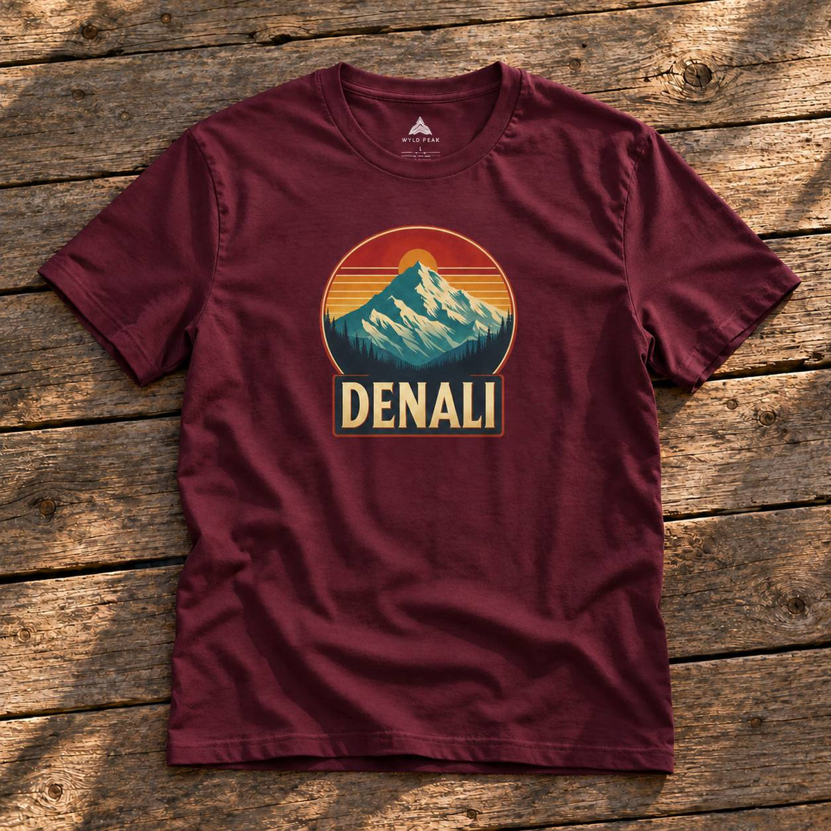 A maroon short-sleeve t-shirt with a retro mountain sunset graphic and the word 'DENALI' printed on the chest, laid flat on a rustic wooden plank surface.