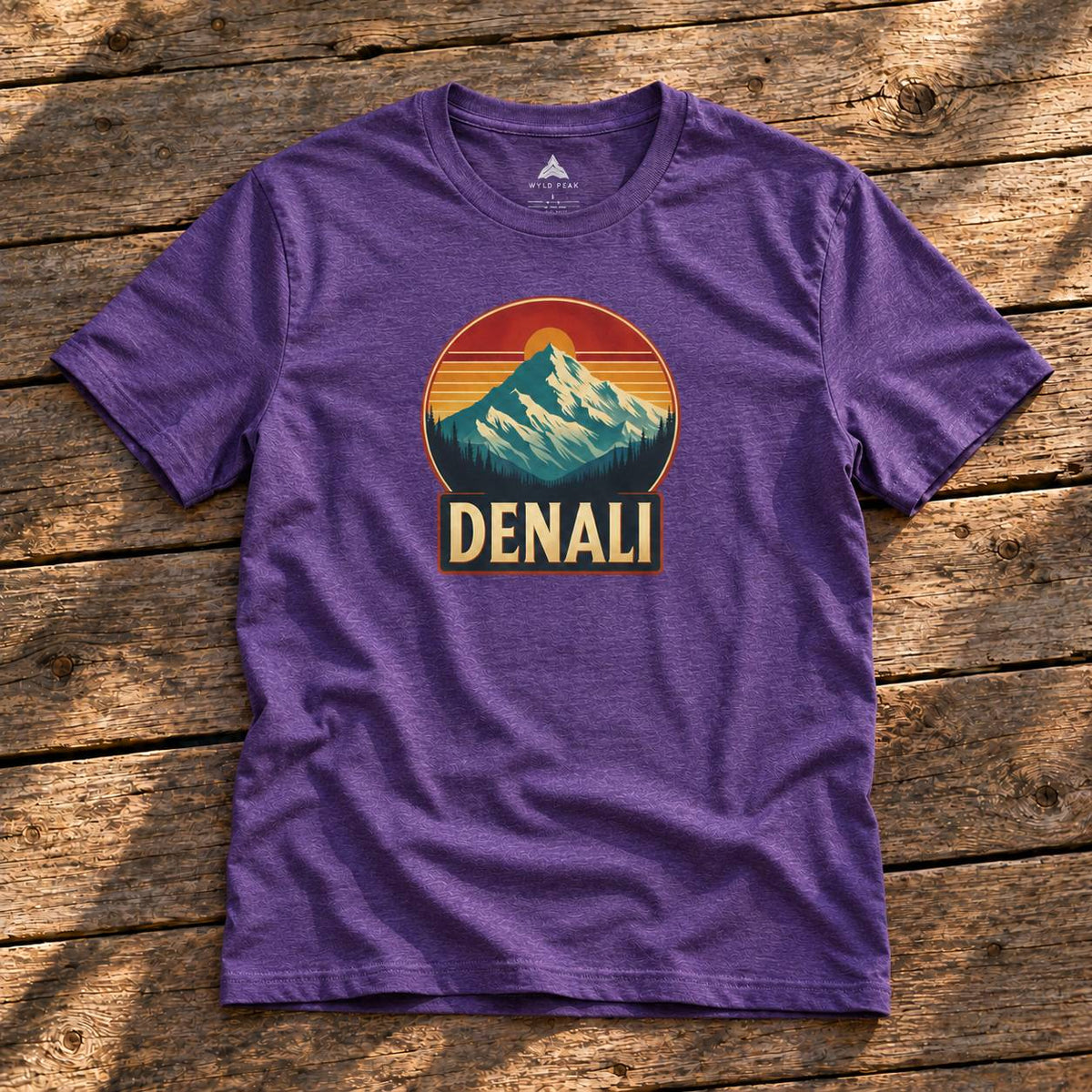 A purple heather short-sleeve t-shirt with a retro mountain sunset graphic and the word 'DENALI' printed on the chest, laid flat on a rustic wooden plank surface.