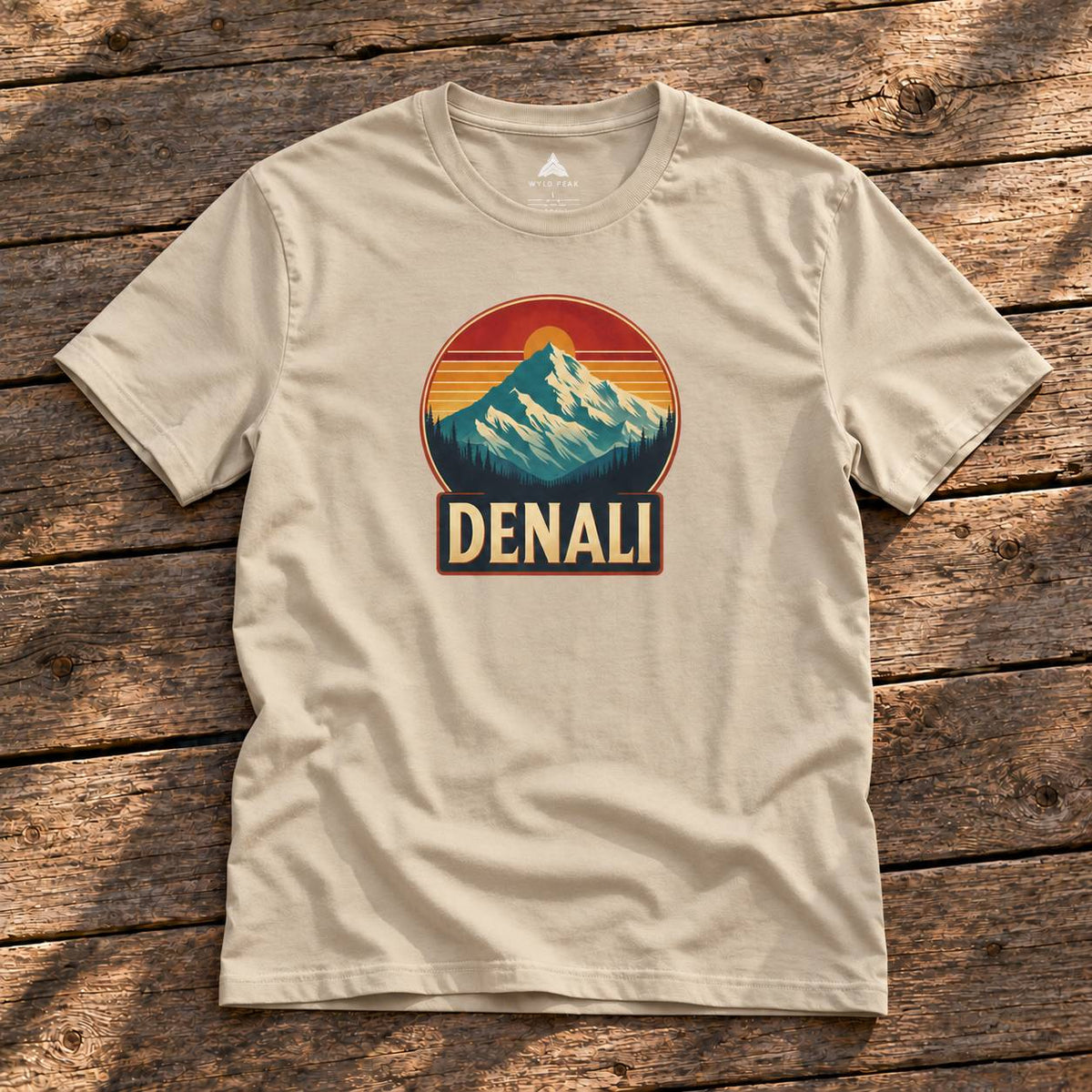 A beige short-sleeve t-shirt laid flat on a rustic wooden surface, featuring a retro-style mountain graphic with a sunset and the word 'DENALI' on the chest.