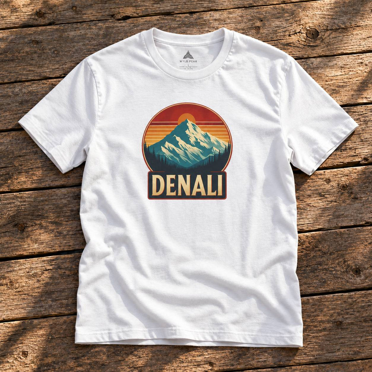 A white short-sleeve t-shirt laid flat on a rustic wooden surface, featuring a retro-style mountain graphic with the word 'DENALI' on the chest.