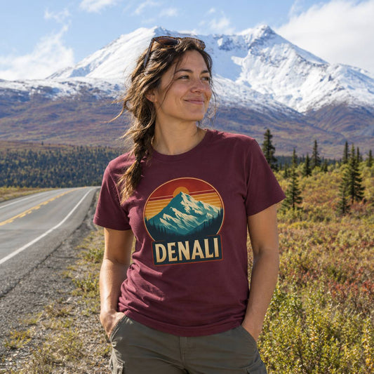 A woman stands outdoors in front of snow-capped mountains and a road, wearing a maroon graphic T-shirt with a mountain-and-sun design and the word "DENALI" on the front.