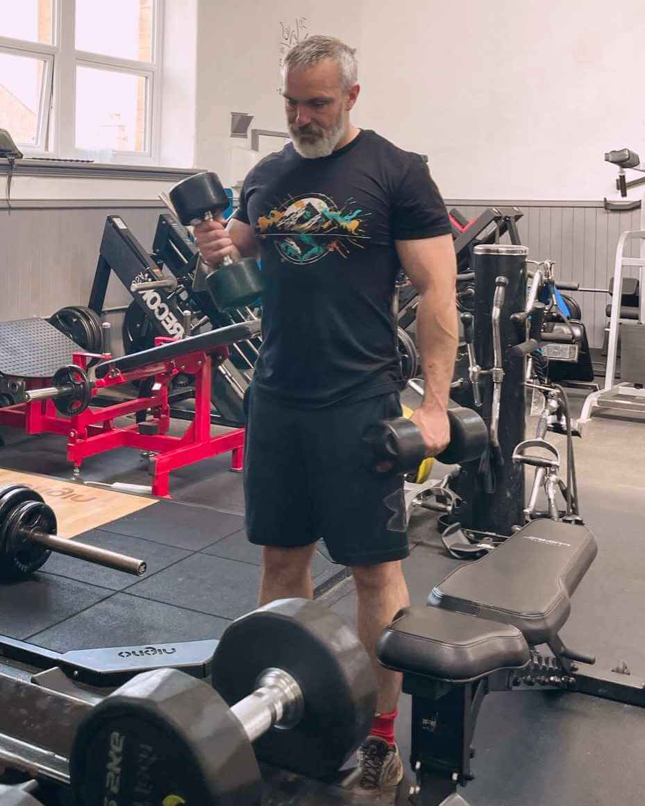 Des in the UK lifting weights at a gym wearing a Wyld Peak organic peak design t-shirt.
