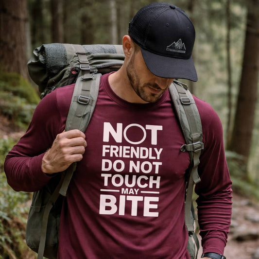 Man in a forest wearing a maroon shirt with a humorous text design and a backpack.