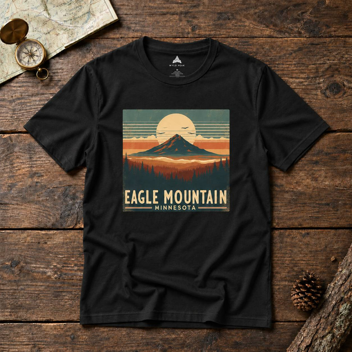 A black short-sleeve graphic T-shirt laid flat on a rustic wooden surface, featuring a retro mountain sunset print with the text 'EAGLE MOUNTAIN MINNESOTA.' Decorative props including a map, compass, and pinecone surround the shirt.