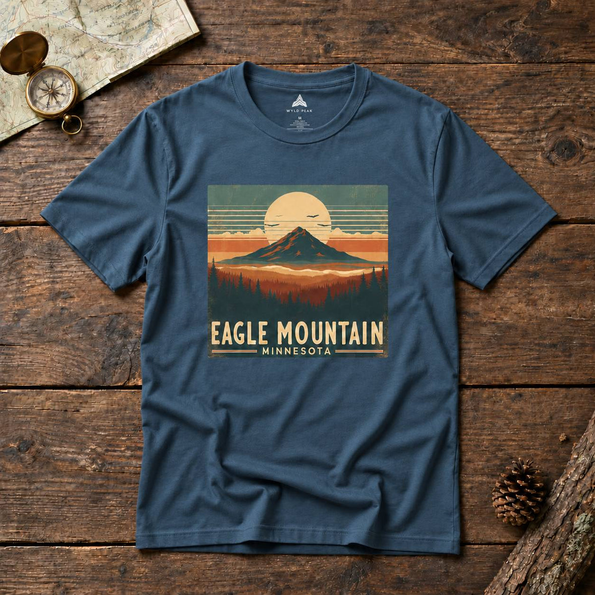 A navy blue short-sleeve t-shirt with a retro-style mountain graphic on the chest reading 'EAGLE MOUNTAIN MINNESOTA'. The shirt is laid flat on a rustic wooden surface with a vintage map, compass, and pinecone nearby.
