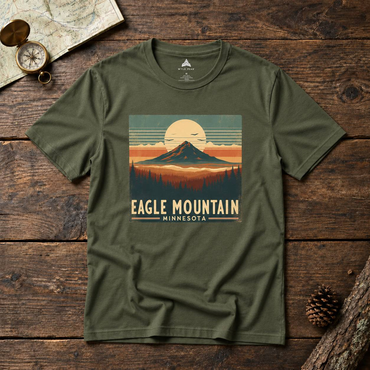 An olive green short-sleeve t-shirt with a vintage-style 'Eagle Mountain Minnesota' graphic print, laid flat on a rustic dark wood background with a map, compass, and pinecone props.
