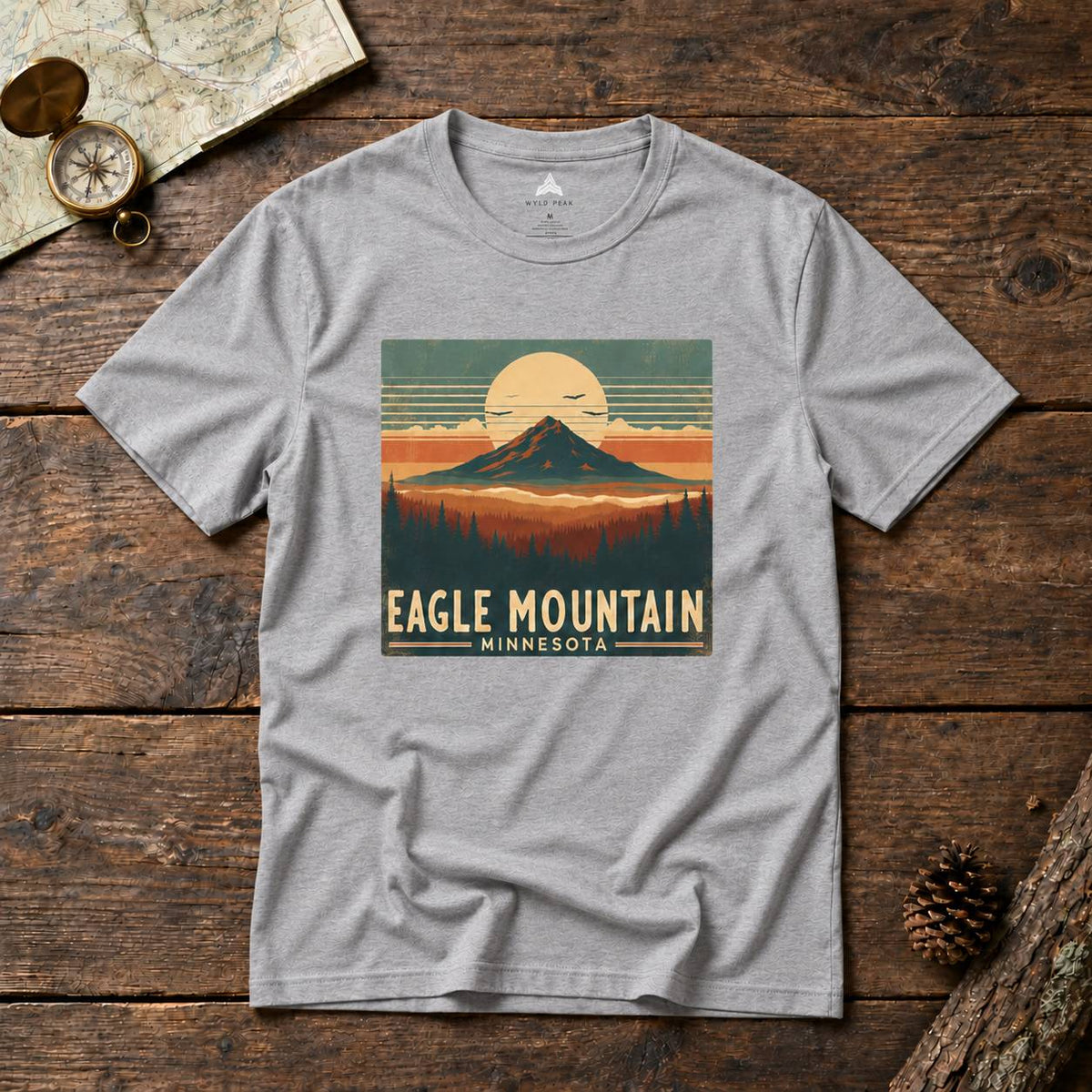 A heather gray short-sleeve t-shirt with a vintage-style 'Eagle Mountain Minnesota' graphic print, laid flat on a rustic dark wood background with a map, compass, and pinecone props.