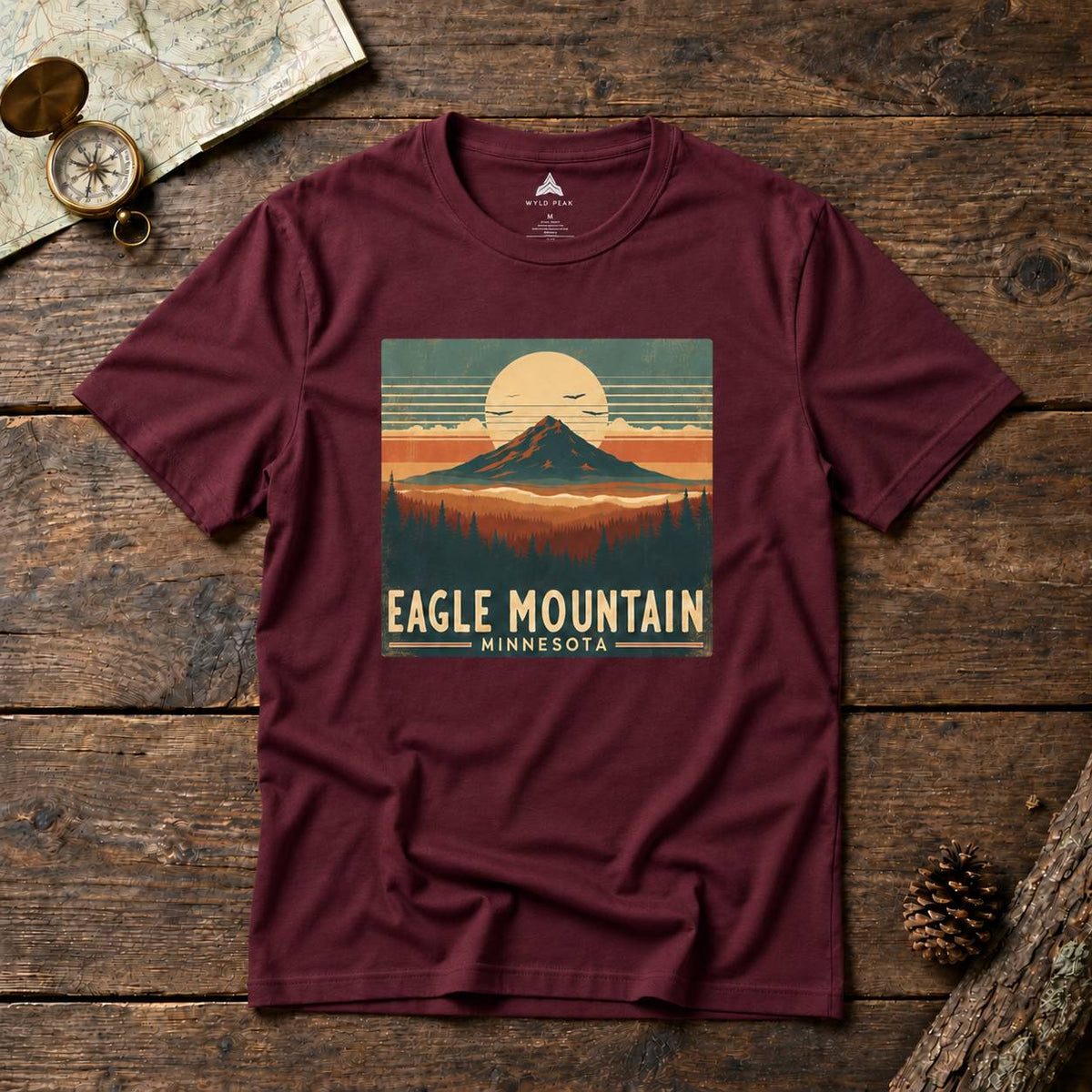 A maroon short-sleeve graphic T-shirt laid flat on a rustic wooden surface, featuring a retro mountain sunset print with the text 'EAGLE MOUNTAIN MINNESOTA.' A map, compass, and pinecone are styled around it as props.