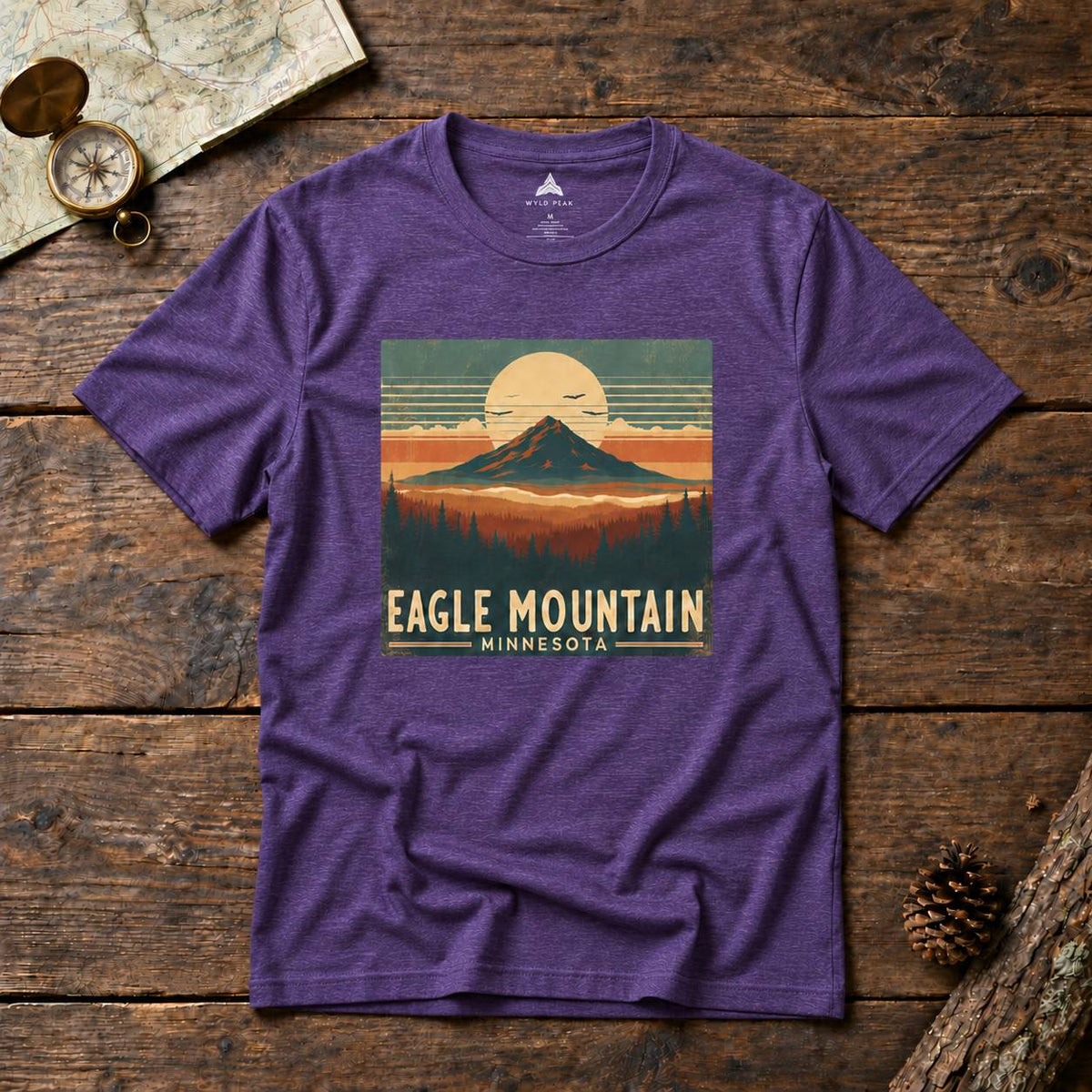 A heather purple short-sleeve t-shirt with a retro-style mountain graphic on the chest reading 'EAGLE MOUNTAIN MINNESOTA'. The shirt is laid flat on a rustic dark wood background with a vintage map, compass, and pinecone as props.