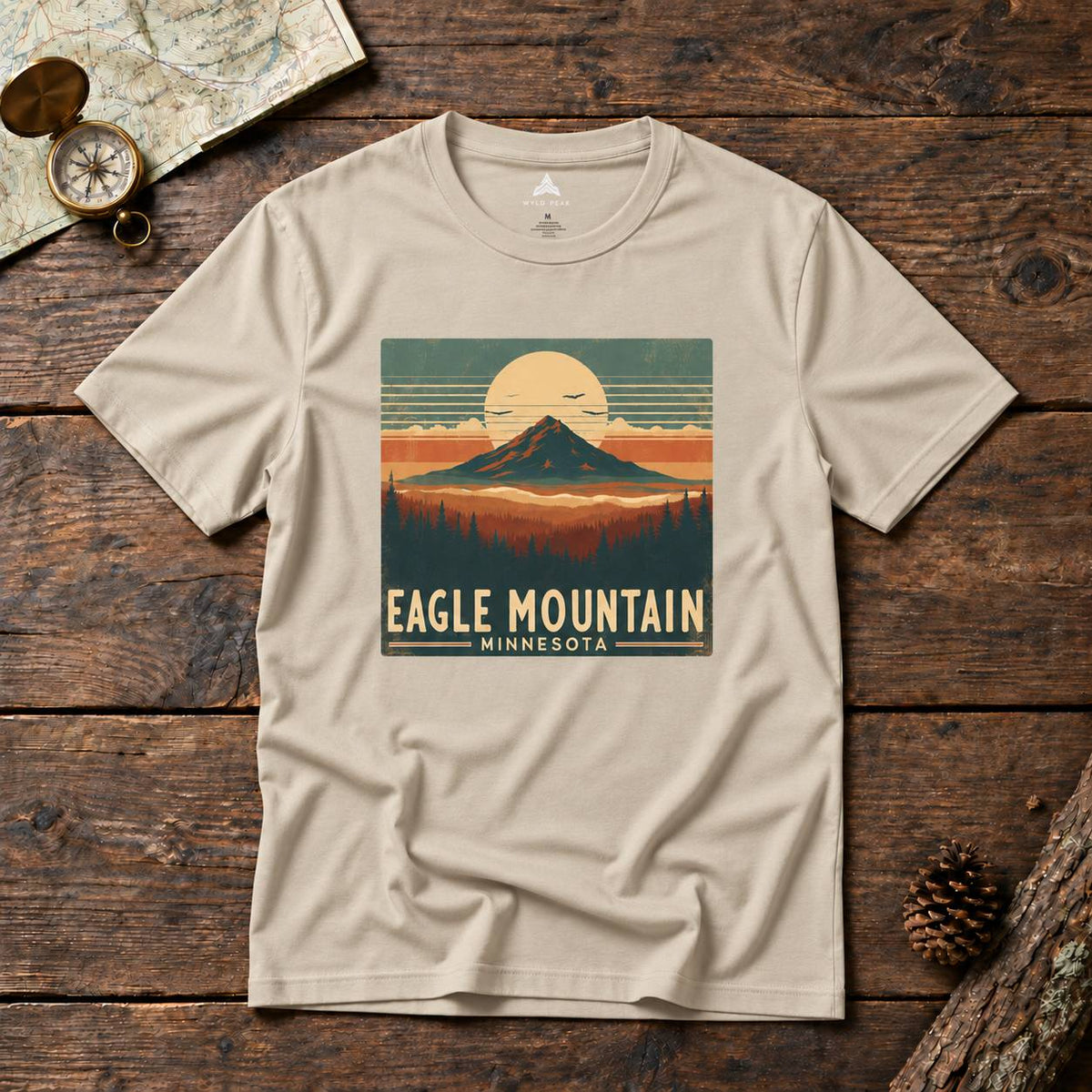 A beige short-sleeve graphic T-shirt laid flat on a rustic dark wood surface, featuring a retro mountain sunset print with the text 'EAGLE MOUNTAIN MINNESOTA.' Decorative props including a map, compass, and pinecone surround the shirt.
