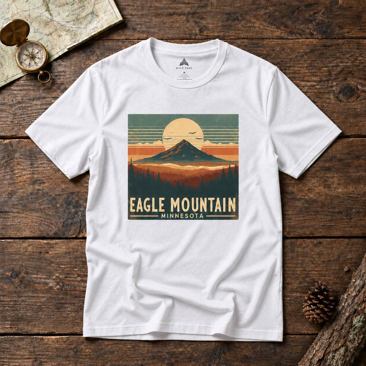 A white short-sleeve t-shirt with a retro-style 'Eagle Mountain Minnesota' graphic print, laid flat on a rustic dark wood background with a map, compass, and pinecone props.