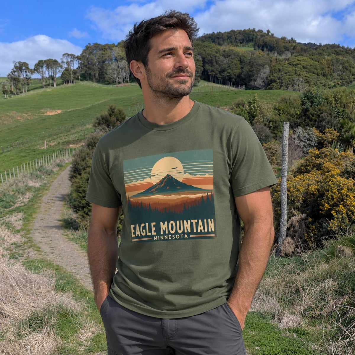 A man wearing an olive green graphic t-shirt with a retro-style 'Eagle Mountain Minnesota' print, standing outdoors on a grassy path with hills and trees in the background.