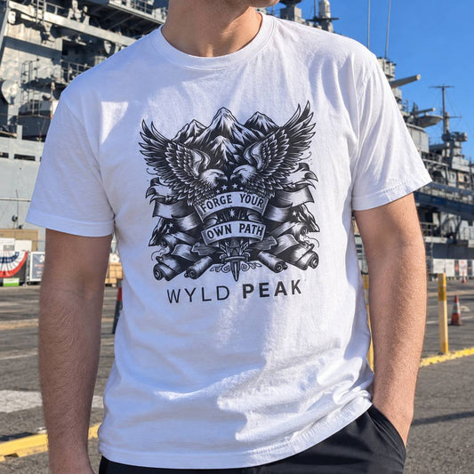 A man wearing a white crewneck t-shirt with a black graphic of two eagles, mountains, and cannons, with the text 'FORGE YOUR OWN PATH' and 'WYLD PEAK'. The background shows a naval vessel dock in daylight.