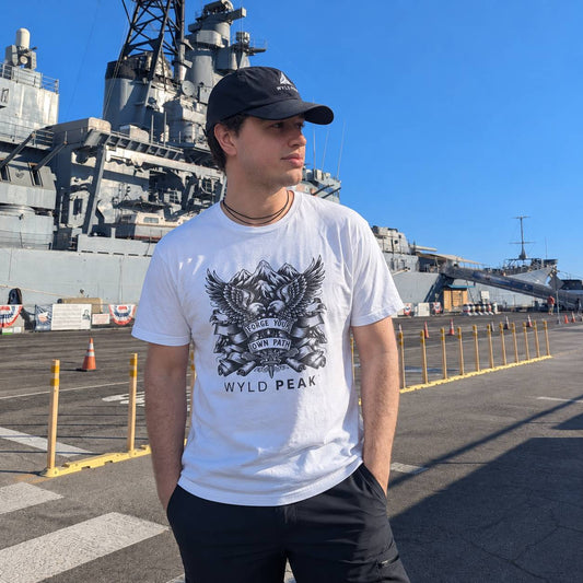 A man wearing a white graphic t-shirt with a black eagle design and the text 'WYLD PEAK' stands on a shipyard deck with a large gray battleship in the background under a clear blue sky.