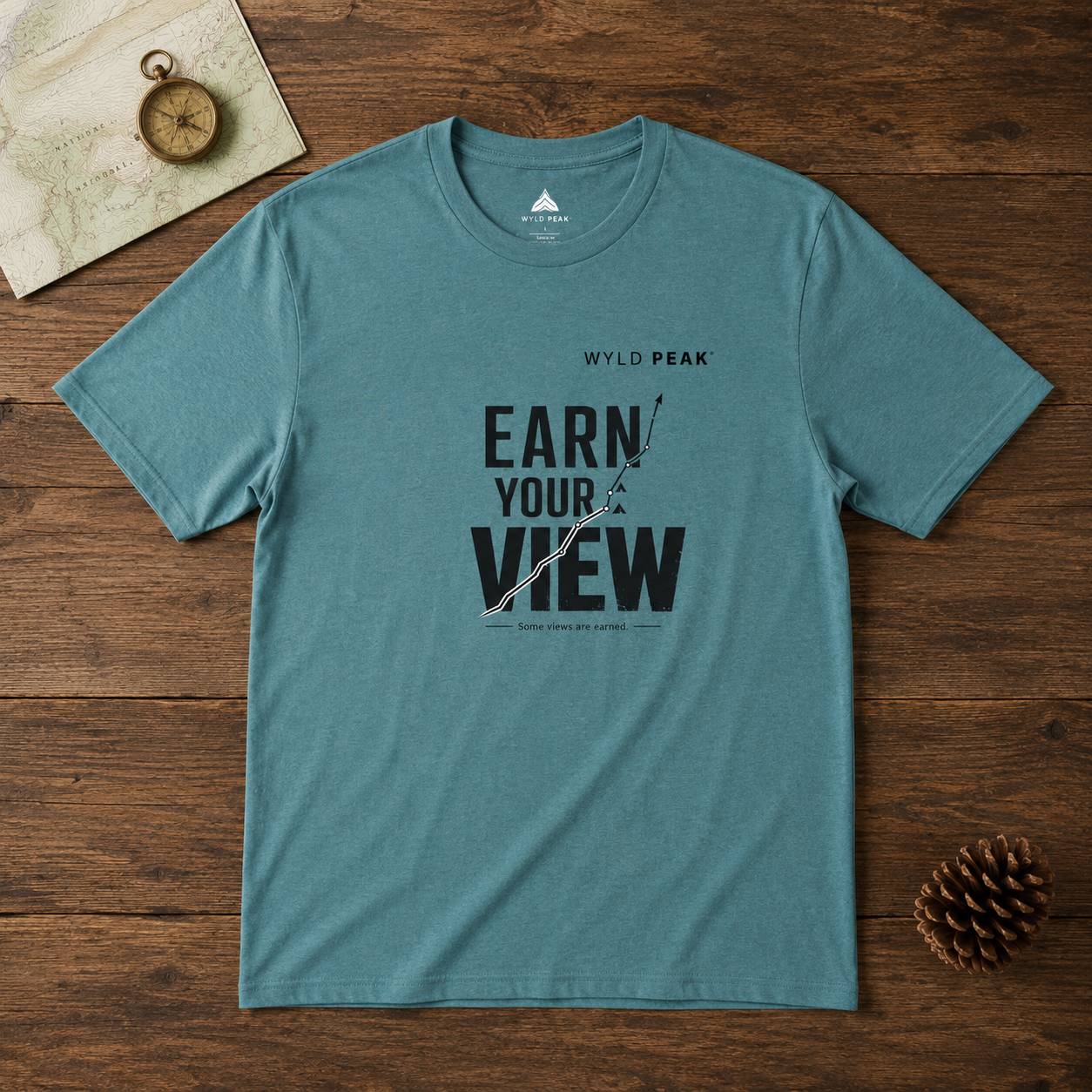 Teal t-shirt with 'Earn Your View' text on a wooden surface with a compass and map.