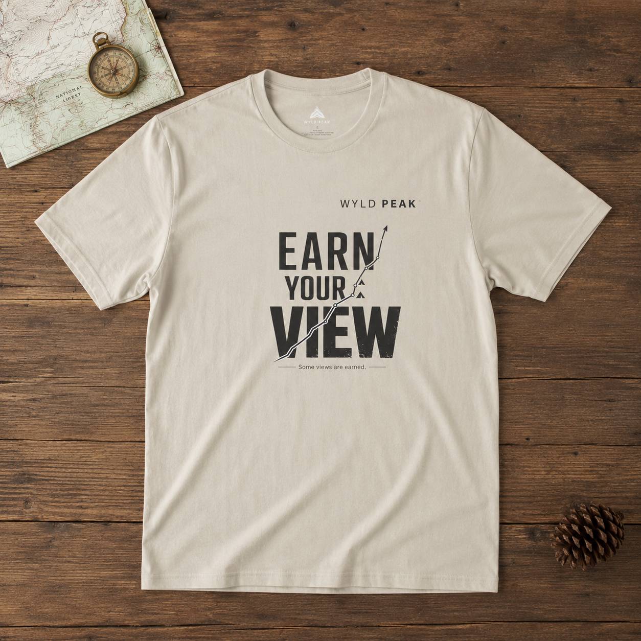 Beige t-shirt with 'Earn Your View' text on a wooden surface with map and compass.
