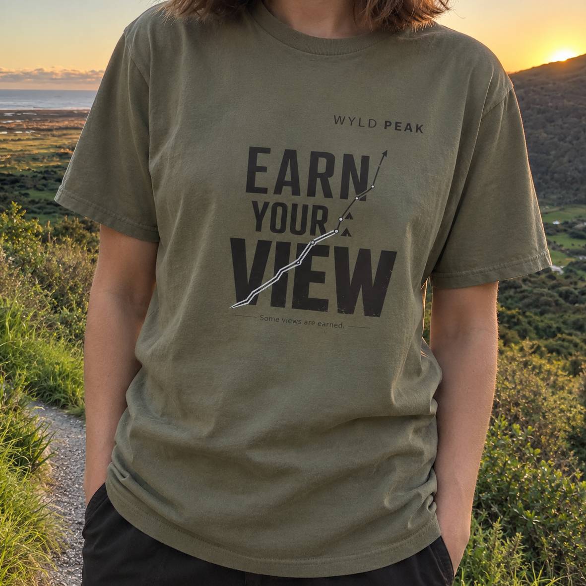 A person outdoors wearing an olive green graphic T-shirt with the text 'EARN YOUR VIEW' and 'WYLD PEAK' printed on the front, with a scenic landscape in the background at sunset.