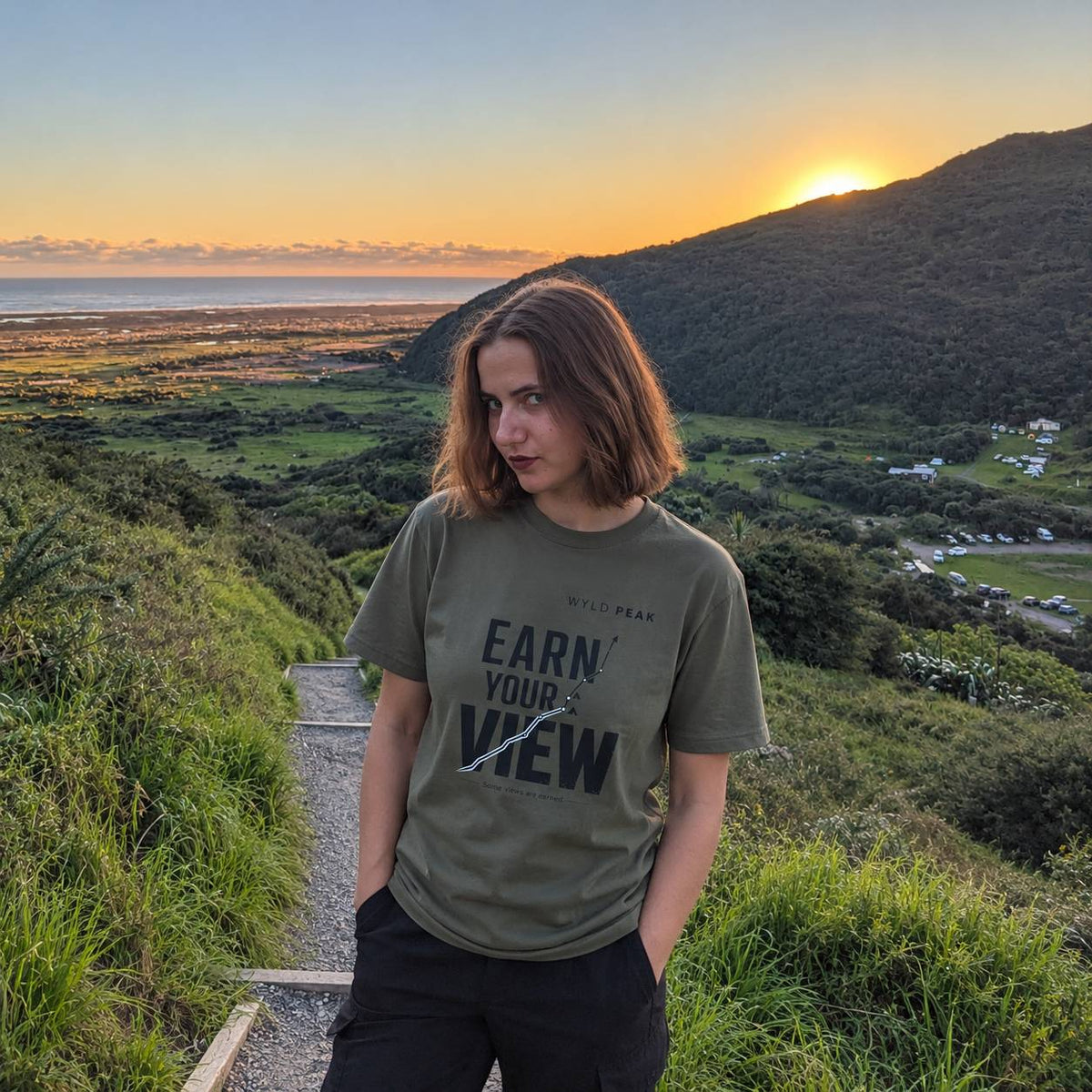 A woman stands outdoors on a hillside path at sunset wearing an olive green graphic T-shirt with the text 'EARN YOUR VIEW' and 'WYLD PEAK' printed on the front, paired with black pants. The background shows a scenic landscape with a valley, ocean, and mountains during golden hour.